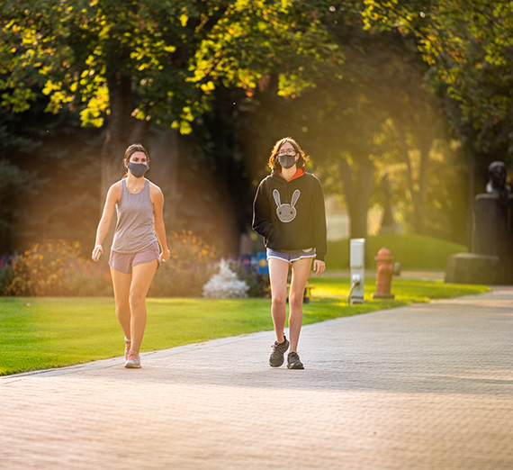 Two students wearing masks walk along a path on campus.