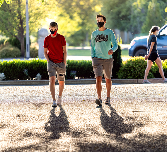 Two students walk through campus in the summertime while wearing masks. 