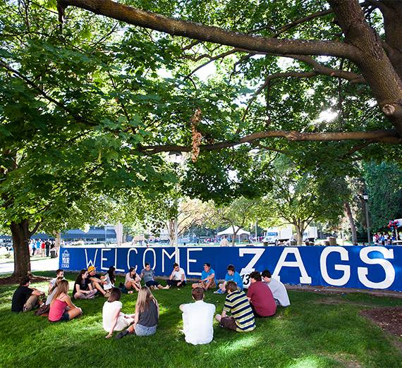 New students sit in a circle on the grass in front of a sign welcoming them to campus. 