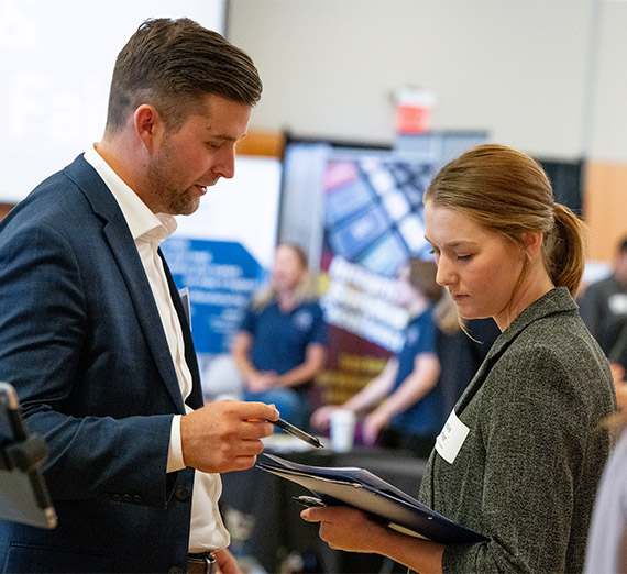 A man and a woman look at the woman's folder and have a conversation during a career fair.  