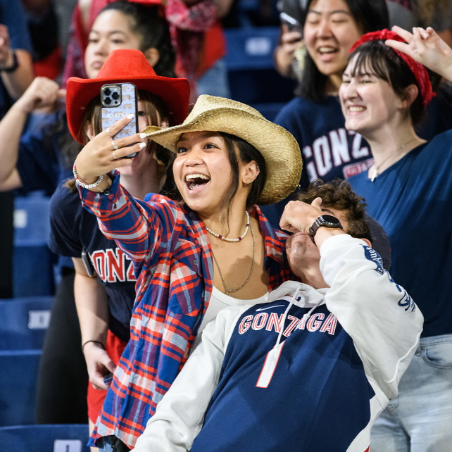 Four students in the Kennel's student section taking a selfie together with a phone. The student holding the phone is smiling and wearing a straw cowboy hat, white shirt, and a red and blue plaid shirt. The three other students are also wearing red, white, and blue clothing.