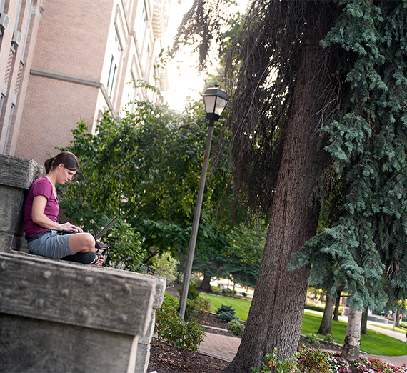 A student sits on a stone bench and works on her laptop. 