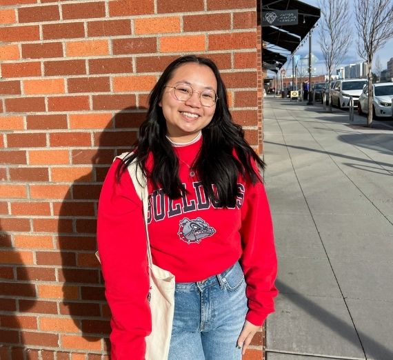 A photo of a current student in a red Gonzaga crewneck sweater smiling. She is holding a white tote bag. She is outdoors, in front of a brick building. 