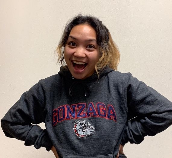 A photo of a current Gonzaga student smiling. She is wearing a gray sweatshirt that spells GONZAGA in navy letters outlined by red. There is a bulldog also on the sweatshirt. 
