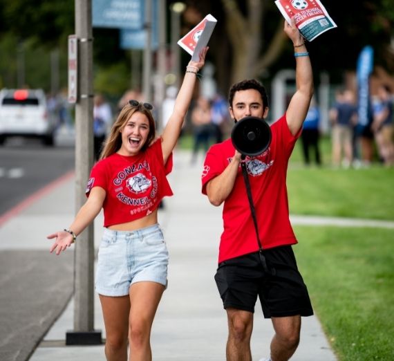 Two current students in red Gonzaga shirts walking down a sidewalk. The student on the left has her right arm in the air with a big smile. The student on the right also has his right arm up and has a bullhorn up to his mouth. 