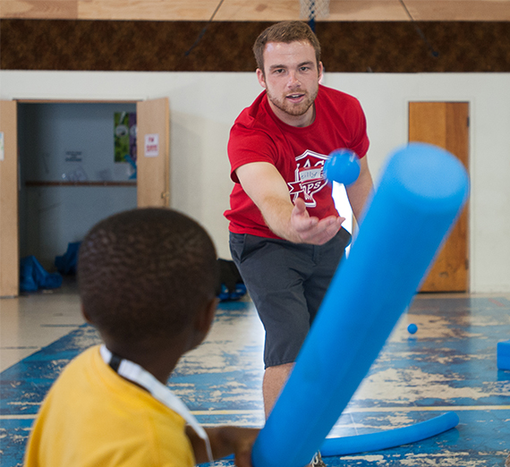 A student volunteer pitches a ball to a student using a pool noodle as a baseball bat.