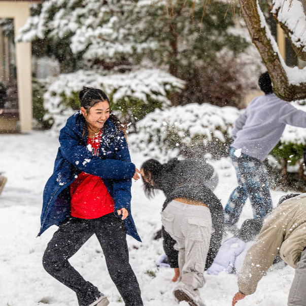 On the left, girl in a red 'Gonzaga' tee shirt throws a snowball at another person.  In the background, students are playing in the snow for a snowball fight.