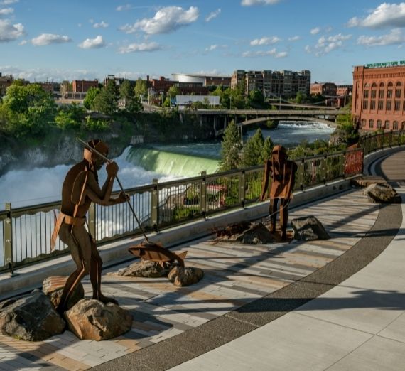 A photo from Riverfront Park overlooking the river. Metal statues line the bridge. In the background, there are waterfalls in the river.