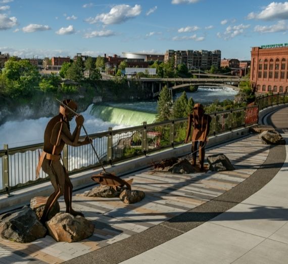 A photo from Riverfront Park overlooking the river. Metal statues line the bridge. In the background, there are waterfalls in the river. 