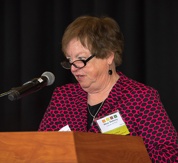 Woman with glasses and short, brown hair speaks at a podium in front of a black background.