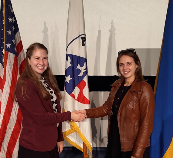 Two women smiling and shaking hands in front of three flags