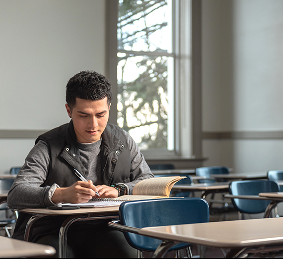 Student sits at a desk in a classroom with an open book on the desk.