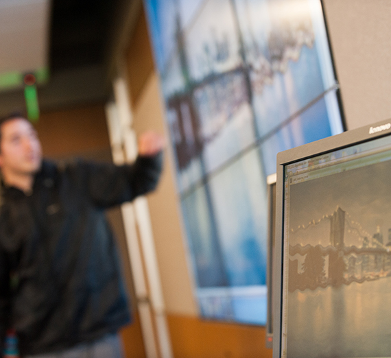 Computer screen in foreground. Student stands in front of large screen in background showing same image.