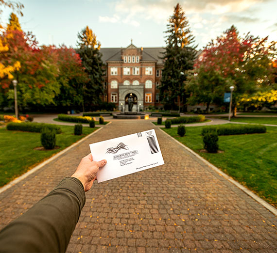 Person holding a vote by mail envelope in front of College Hall surrounded by trees in autumn.