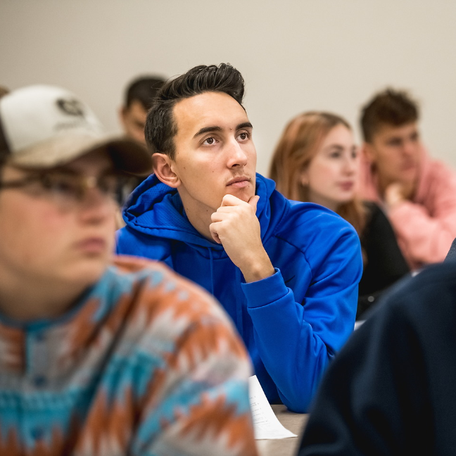 A photo of a classroom with many students. The photo is focused on a male student sitting in the center of the classroom. The student has black hair and is wearing a bright blue sweatshirt. He is leaning on his arm with his chin resting on his hand. The student is looking up attentive and listening. In the foreground, is a student wearing glasses wearing a grey hat and an orange, blue, and grey pullover jacket. There are another three students in the background also listening in the classroom. 
