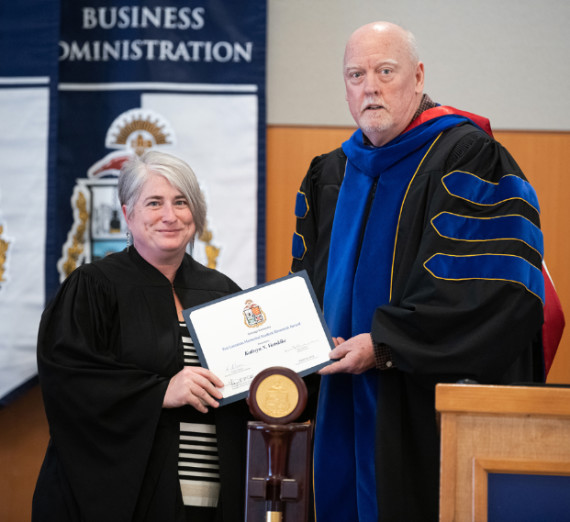 Man hands woman award, both wear academic regalia