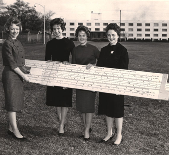 Black and white photo of four women holding a giant slide-rule