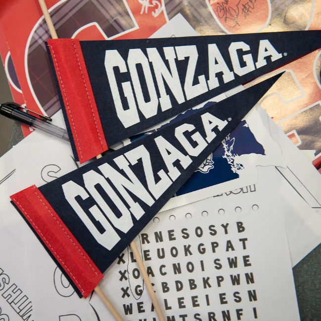 Two Gonzaga pennants one above the other in the picture. The two pennants have a red lining on the left-hand side and are navy blue with white letters spelling out "Gonzaga."  Both pennants have a small wooden stick sticking out of the red lining on the side. The two pennants are sitting on top of various papers including another GU sign that is red with navy plaid letter spelling out "GU." 