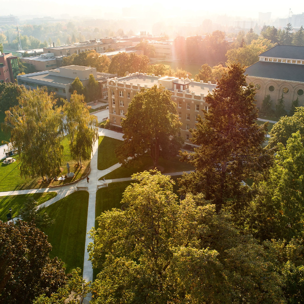 A drone photo overlooking the center of Gonzaga University campus. It shows six pathways in the center of the photo meeting at an Aluminum Jesus statue. Students are walking along these pathways. The photo also overlooks Desmet Hall which is a residence hall. This hall has beige brick with white trim along the building. On the upper right side of the photo is an overlooking view of Magnuson Theatre which has beige and red brick with circular windows on the side of it. There is lots of greenery and trees in the photo.