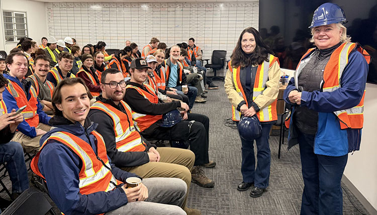 Many young adults sitting in rows wearing safety vests with two women on the right of the frame
