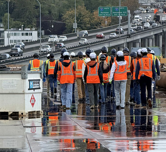 Two dozen people stand together near a freeway wearing safety gear