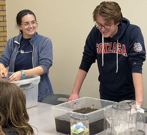 Two students stand at a table holding soil and water in a clear plastic bin