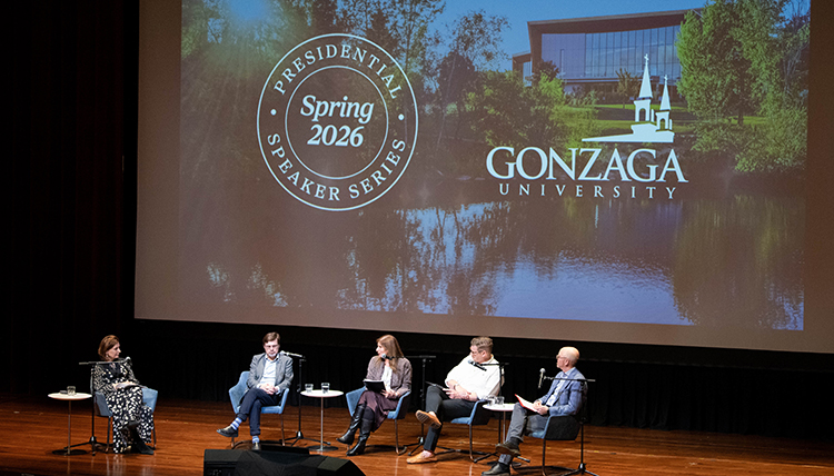 Five people on stage sit in chairs in front of a large screen