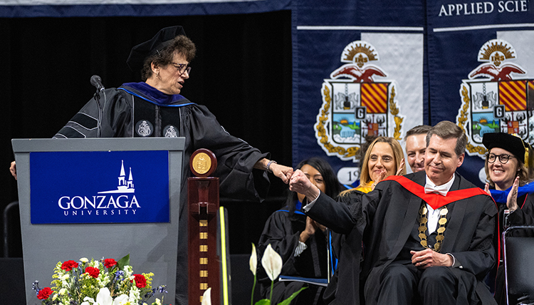 A woman at a podium fist bumps a man sitting in a chair