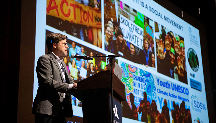A man stands at a podium in front of a screen with several photos