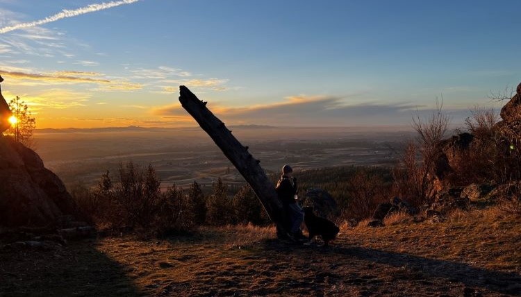 A boy and a dog sitting watching the sun rise from a hiking trail in Spokane, WA