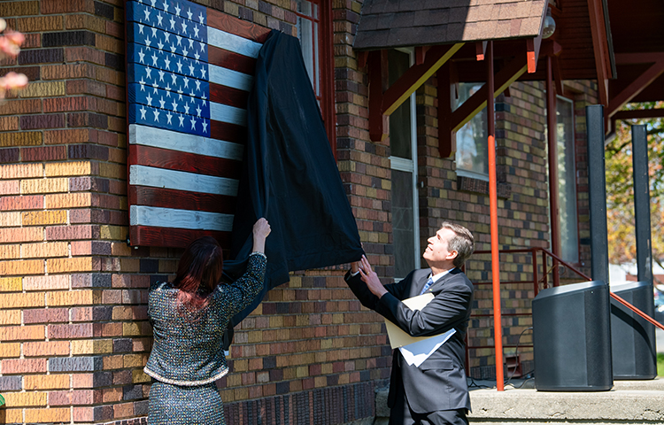 A man and women reveal a flag memorial hanging on the wall of a house.