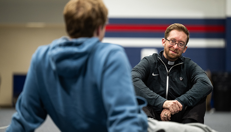 Fr. Tim Breen sits on the floor talking to a student.