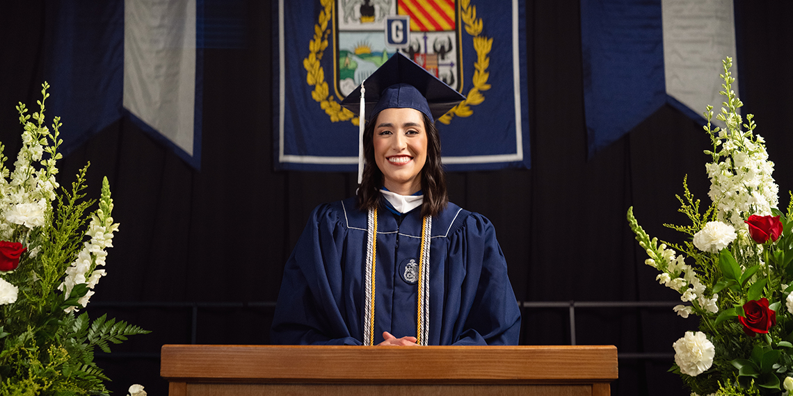 A woman in cap and gown smiles at the camera as she stands between two flower arrangements behind a podium