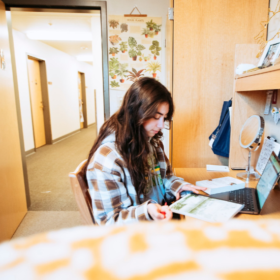 Student sitting at their wooden desk in a dorm. The female student has long dark brown wavy hair and is wearing a brown and white plaid shirt with a grey sweatshirt underneath. The student is looking at a notebook and has a laptop and book in front of her on the desk. In the background is a poster with green plants and then her dorm room door open showing a hallway with grey carpet for the residence hall.