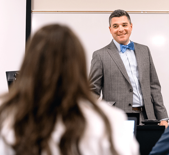 A man in a bow tie speaks to a class as he stands in front of a whiteboard