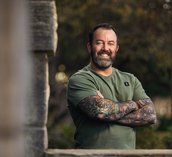 A man with a beard and tattooed arms stands with his arms crossed