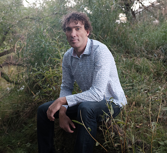 A man with curly brown hair crouches in some brush