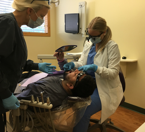 A dentist patient looking at his teeth in a mirror, while a dentist looks over him.