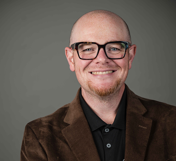 A smiling man with glasses and a goatee in a brown suit jacket