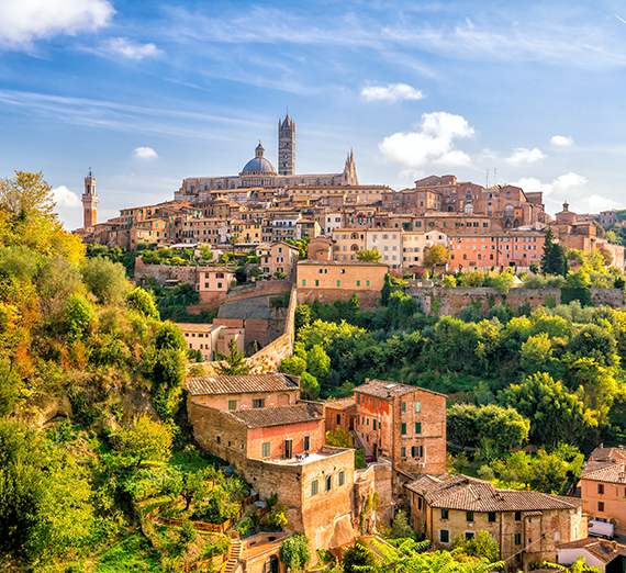 A sunlit view of a hilltop town in Tuscany, Italy, featuring terracotta rooftops, stone buildings, and lush greenery cascading down the hillside. In the background, the majestic Siena Cathedral and its bell tower rise above the city, standing out against a bright blue sky dotted with soft clouds — a perfect blend of medieval architecture and Italian countryside charm.