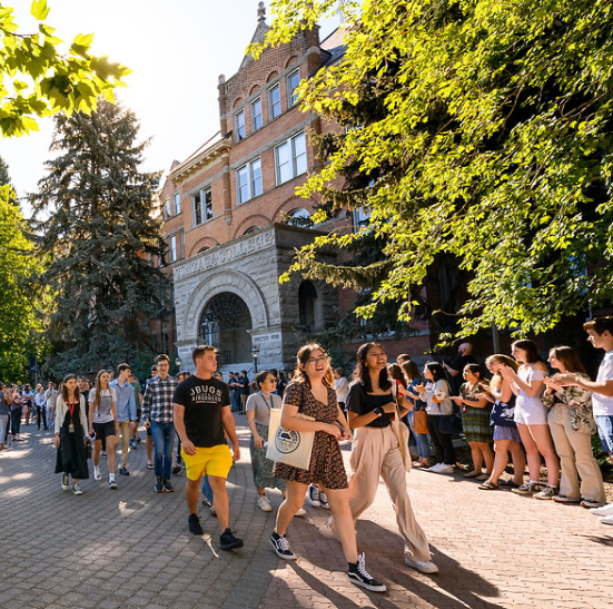 A group of students walk along a brick pathway in front of a large stone and brick building on campus, College Hall. There are current students, faculty, staff lining the pathway and clapping for the students walking down the middle of the pathway. these students walking down the pathway are new students on their way to Academic Convocation. The two students in the forefront are both young women leading the group of students walking. The young woman on the left is wearing a black dress with orange flowers. The young woman to her right is wearing light beige pants and a black blouse. Students are smiling as they walk along the path. 