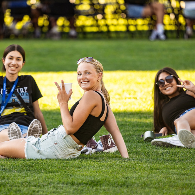 Three students sitting on grass smiling toward the camera. In the background, there are students sitting at tables for orientation.