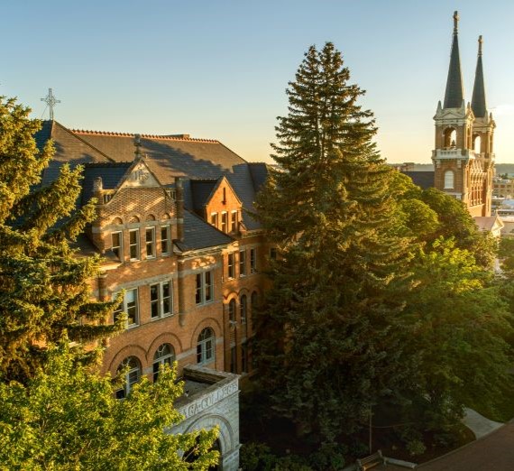 The front of College Hall from an aerial perspective. The building has a stone facing and is red brick. There are trees in front of the building with the sun shining thorugh and the spires of a church in the background.
