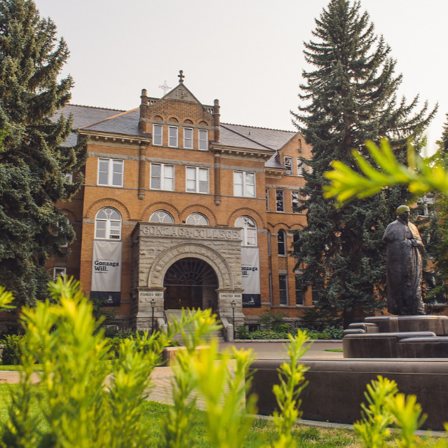 College Hall as a red-brick building with a stone front entryway. It is surrounded by green foliage and in front of the building is a statue of St. Ignatius.