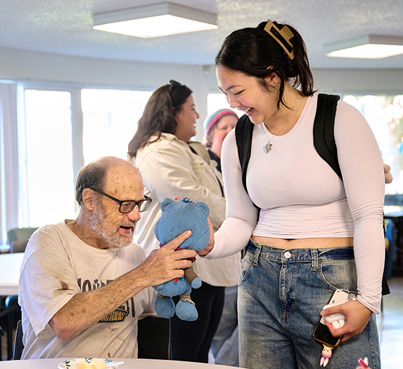 A ������ volunteer smiles while handing a stuffed toy to a senior community member during a service event, creating a joyful moment of connection.