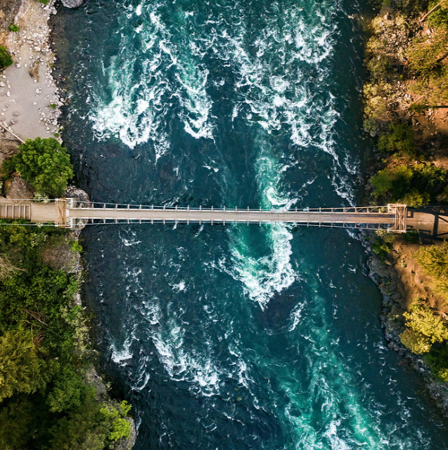 Overhead drone shot of the Spokane River flowing underneath a wooden bridge. On either side of the river, there is beautiful nature and trees on the banks.