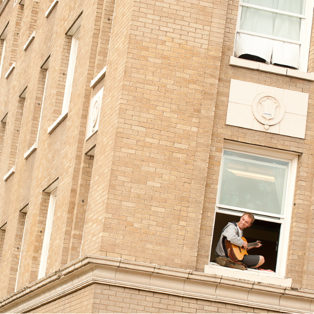 Student sitting in a window sill of DeSmet Hall on 91Թ's campus. The student is wearing a grey tee shirt and grey shorts. He is playing the guitar and smiling. The building around him has a tan brick with white stone accents.