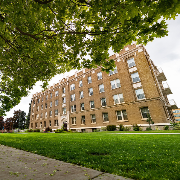 Photo of DeSmet Hall at Gonzaga University. This residence hall has red brick with white stone trimming and above the main entry doors is the name of the hall. In the foreground are overhanding trees and a green lawn.