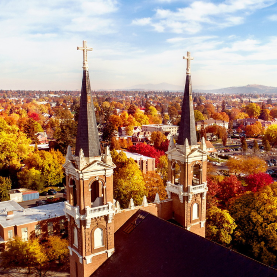 The spires of St. Al's Church in the foreground during the day with a beautiful background of campus with the leaves changing color in autumn.