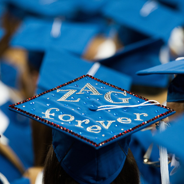 Top of a blue graduation hat with a white tassel amongst a crowd of students wearing graduation hats. The graduation cap is decorated with red gems outlining the hat and then silver gems spelling out "Zag forever."